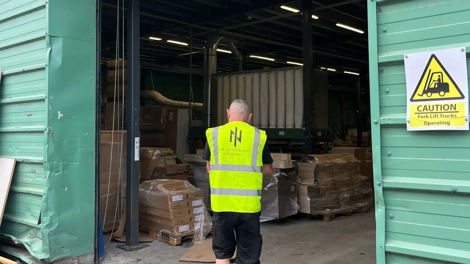 Engineer in NW Automatic Doors hi-vis, seen from behind, walking into a warehouse through an open roller shutter
