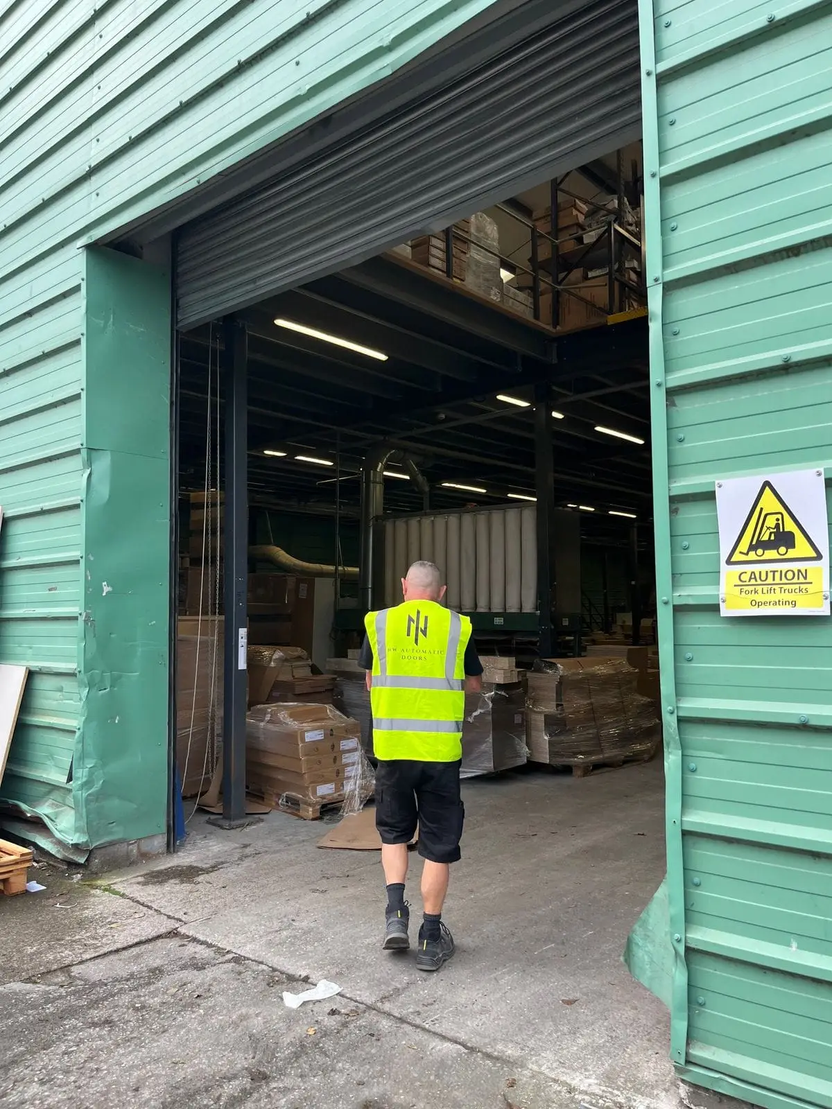 Engineer in hi-vis, seen from behind, walking through an open roller shutter into a warehouse